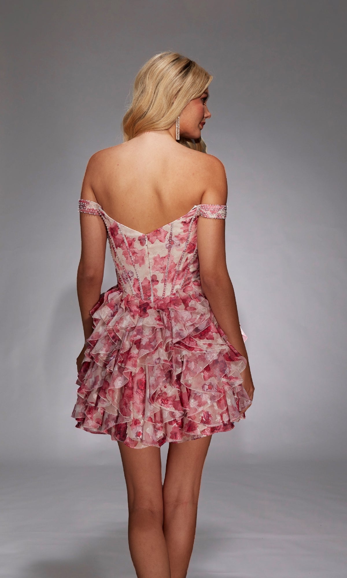 A woman with long blonde hair models the Alyce 30034 short homecoming dress, an off-the-shoulder pink floral fit-and-flare style, shown from the back against a plain gray background.